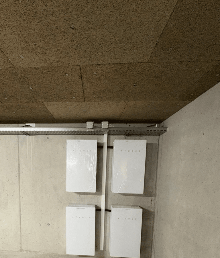 Four white electrical enclosures mounted on a concrete basement wall with metal conduit and a ceiling cable tray above them. | Solution de stockage d’énergie à Yverdon-les-Bains | Nos réalisations