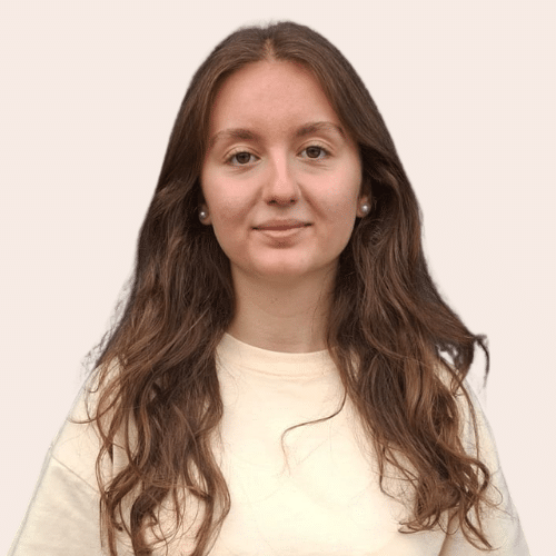 Portrait of a young woman with long wavy brown hair, wearing a light beige sweater, facing the camera with a neutral expression.